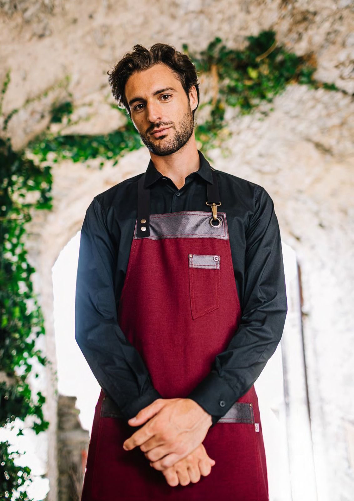 Uomo con camicia nera e grembiule bordeaux in piedi con le mani giunte in un ambiente esterno