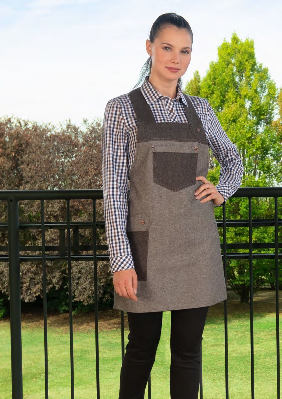 Una donna con una camicia a quadri e un grembiule è in piedi all'aperto, vicino a una ringhiera e a un giardino.