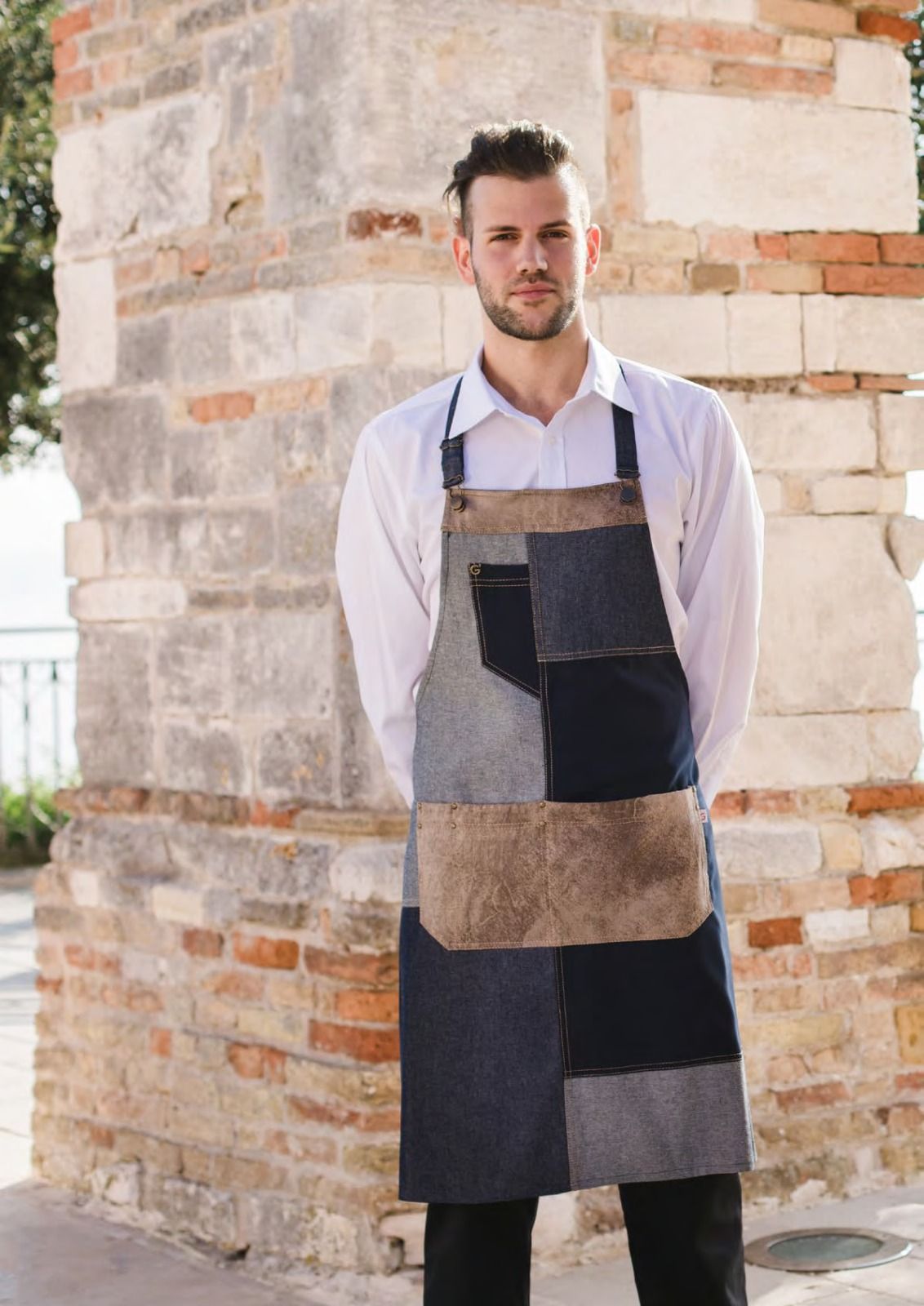 Un uomo con indosso un grembiule a patchwork è in piedi accanto a un pilastro di mattoni all'aperto.