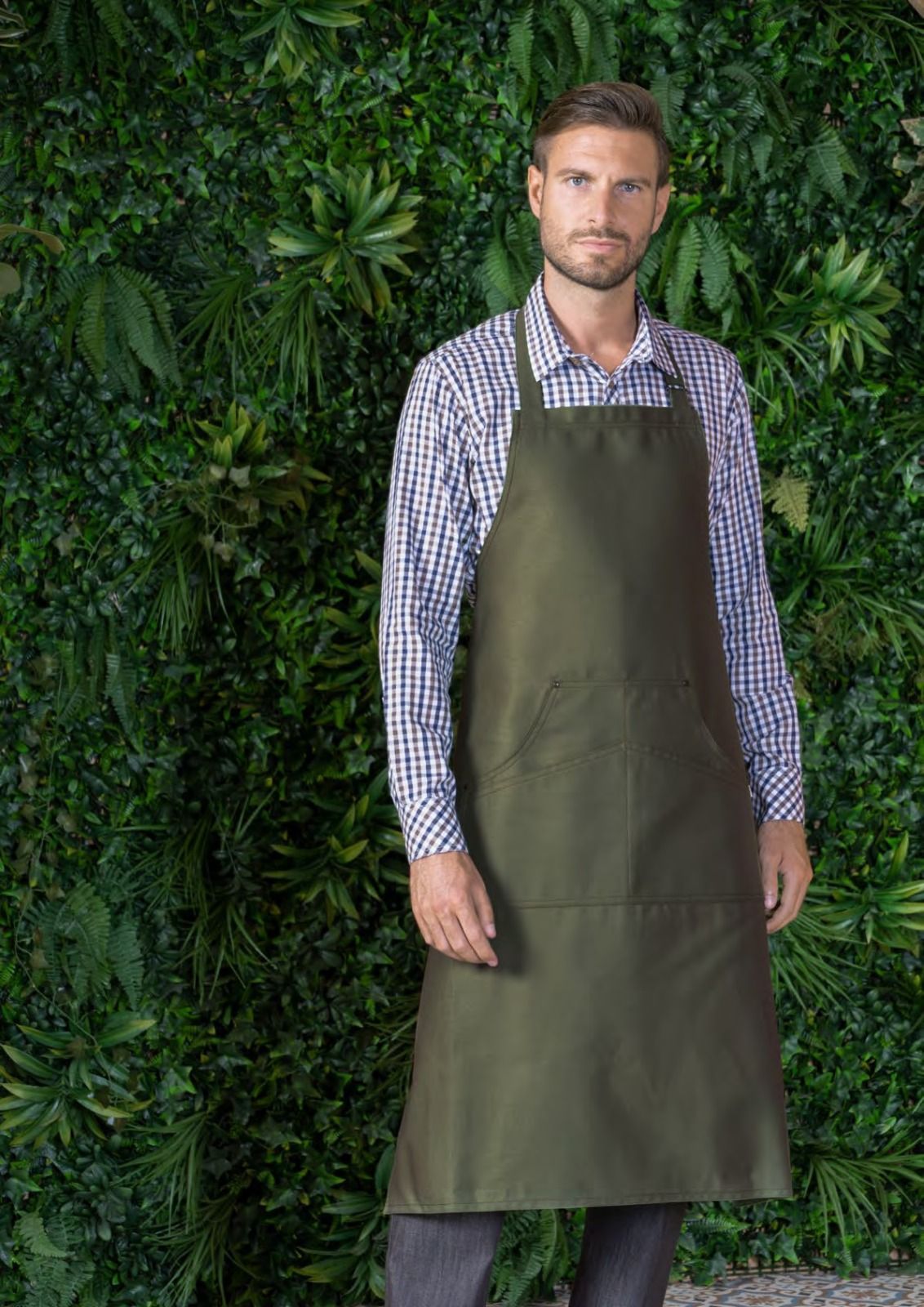 Uomo con camicia a quadri e grembiule verde oliva in piedi davanti a un muro ricoperto di vegetazione verde.