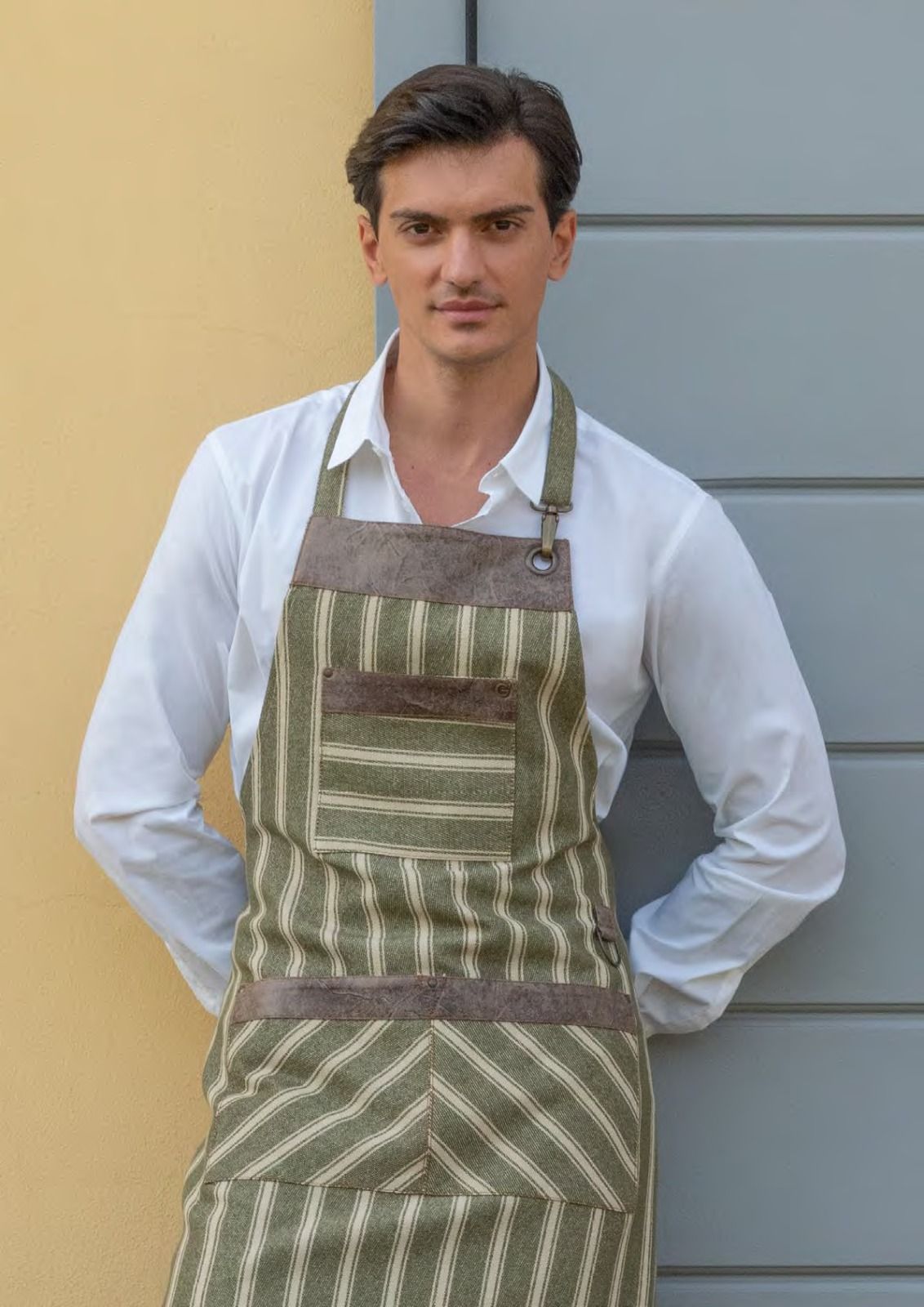 Un uomo con una camicia bianca e un grembiule a righe è in piedi contro un muro grigio, con le mani dietro la schiena.