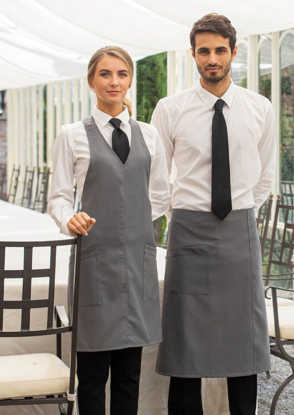Due membri dello staff di un ristorante, con camicie bianche, cravatte nere e grembiuli grigi, sono in piedi nella sala da pranzo.