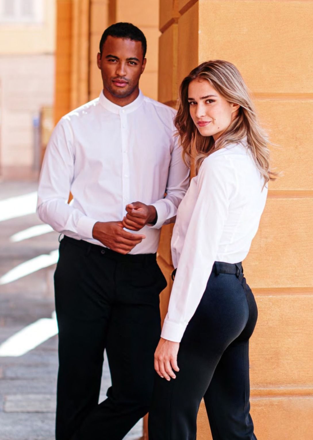 Due persone con camicie bianche e pantaloni neri posano insieme in un corridoio luminoso.
