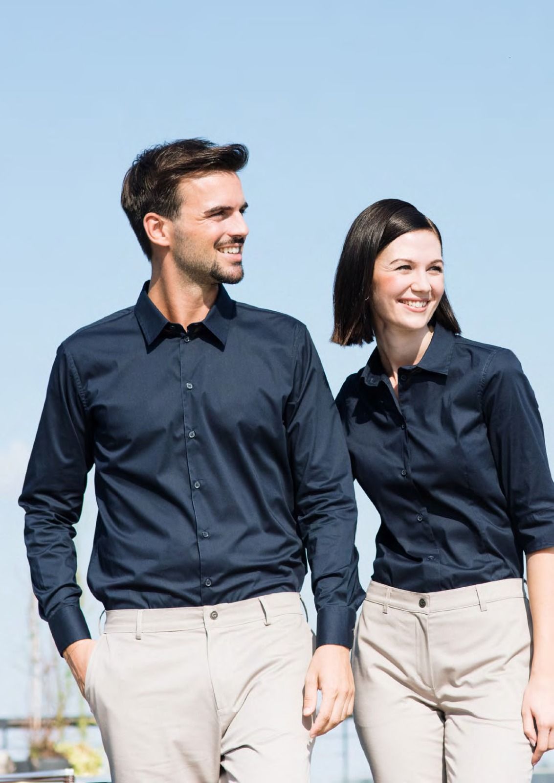 Due persone con camicie blu scuro e pantaloni beige sorridono all'aperto contro un cielo azzurro.