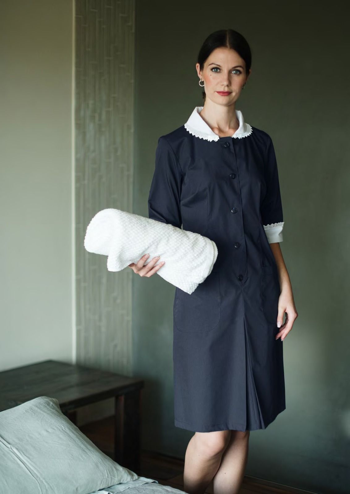 Una donna in abito blu scuro tiene in mano un cuscino bianco in una stanza dai colori tenui.