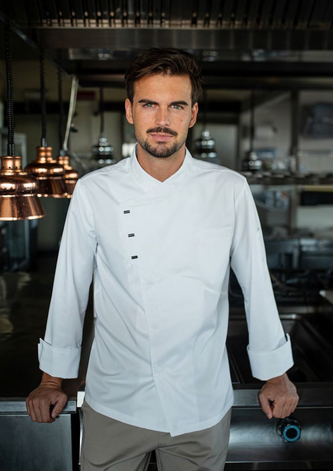 Uno chef in giacca bianca è in piedi nella cucina di un ristorante, appoggiato a un bancone.