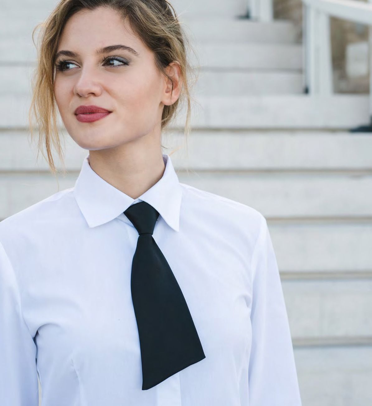 Una donna con camicia bianca e cravatta nera è in piedi su una scalinata e guarda di lato.
