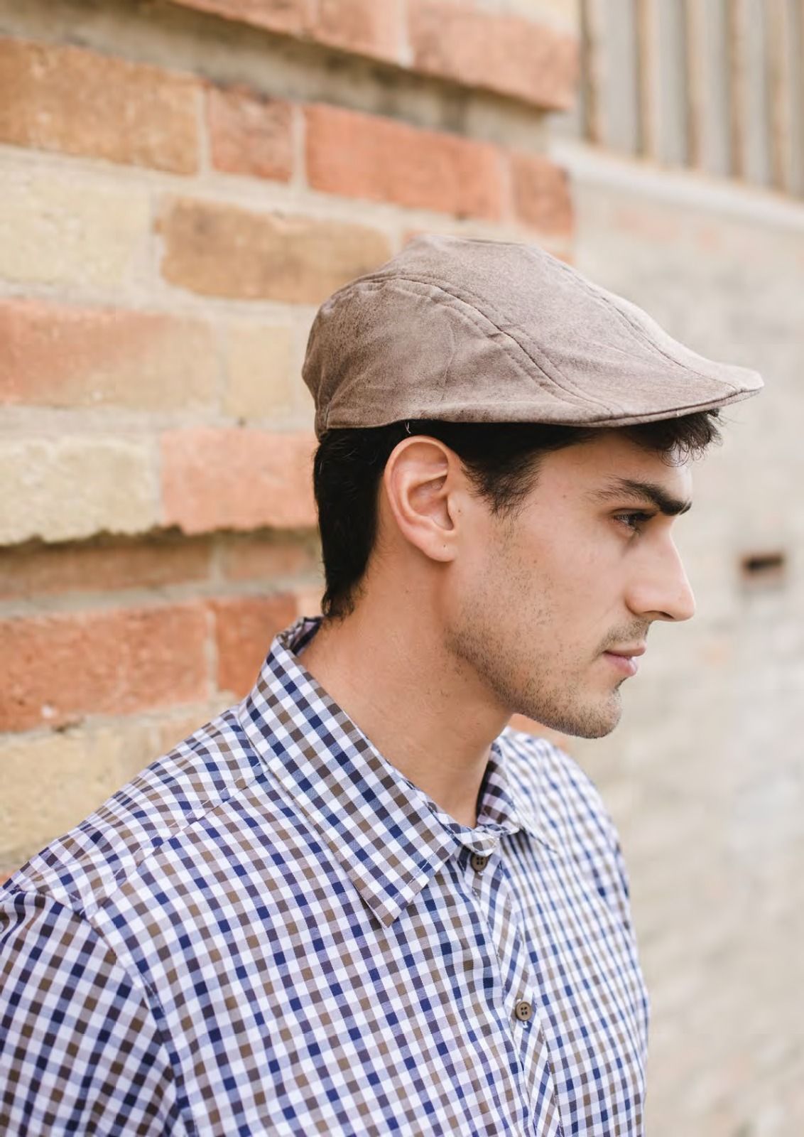 Profilo laterale di una persona con berretto beige e camicia a quadri blu, sullo sfondo di un muro di mattoni.