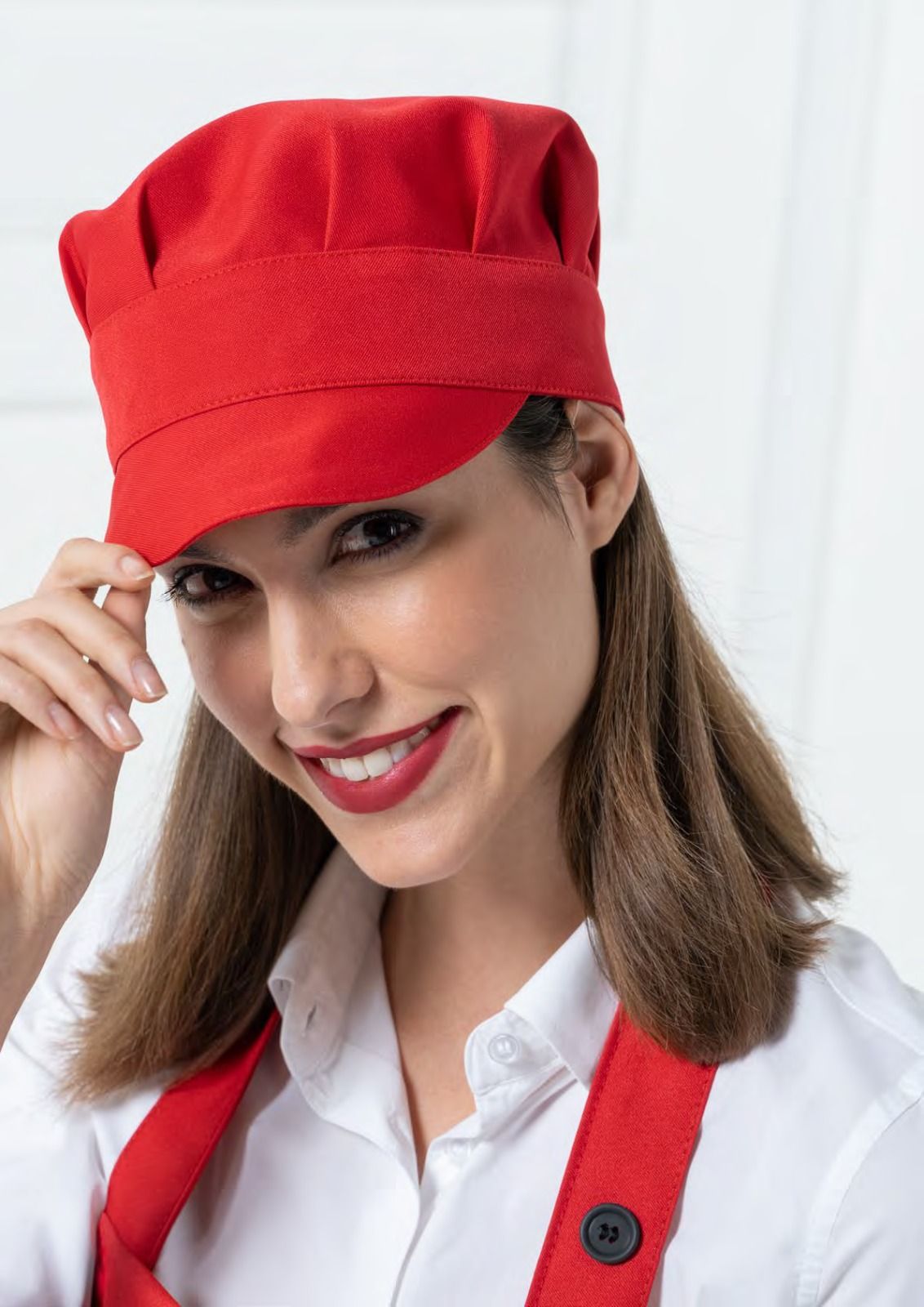 Persona sorridente con cappello e grembiule da chef rossi, che si sistema il cappello su uno sfondo bianco.