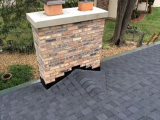 Brick chimney with a damaged base standing on a dark shingled roof