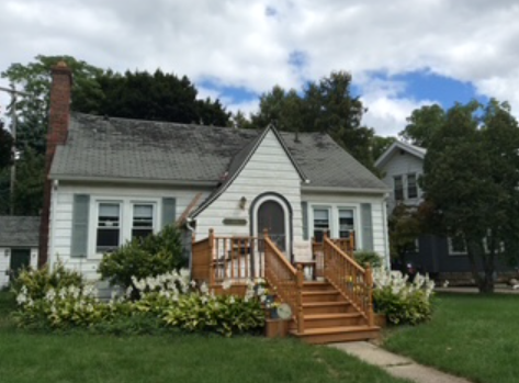 Small white house with gray roof, front porch stairs, and a flower garden in front