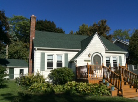 White house with green roof, front porch, and wooden steps surrounded by trees and shrubs