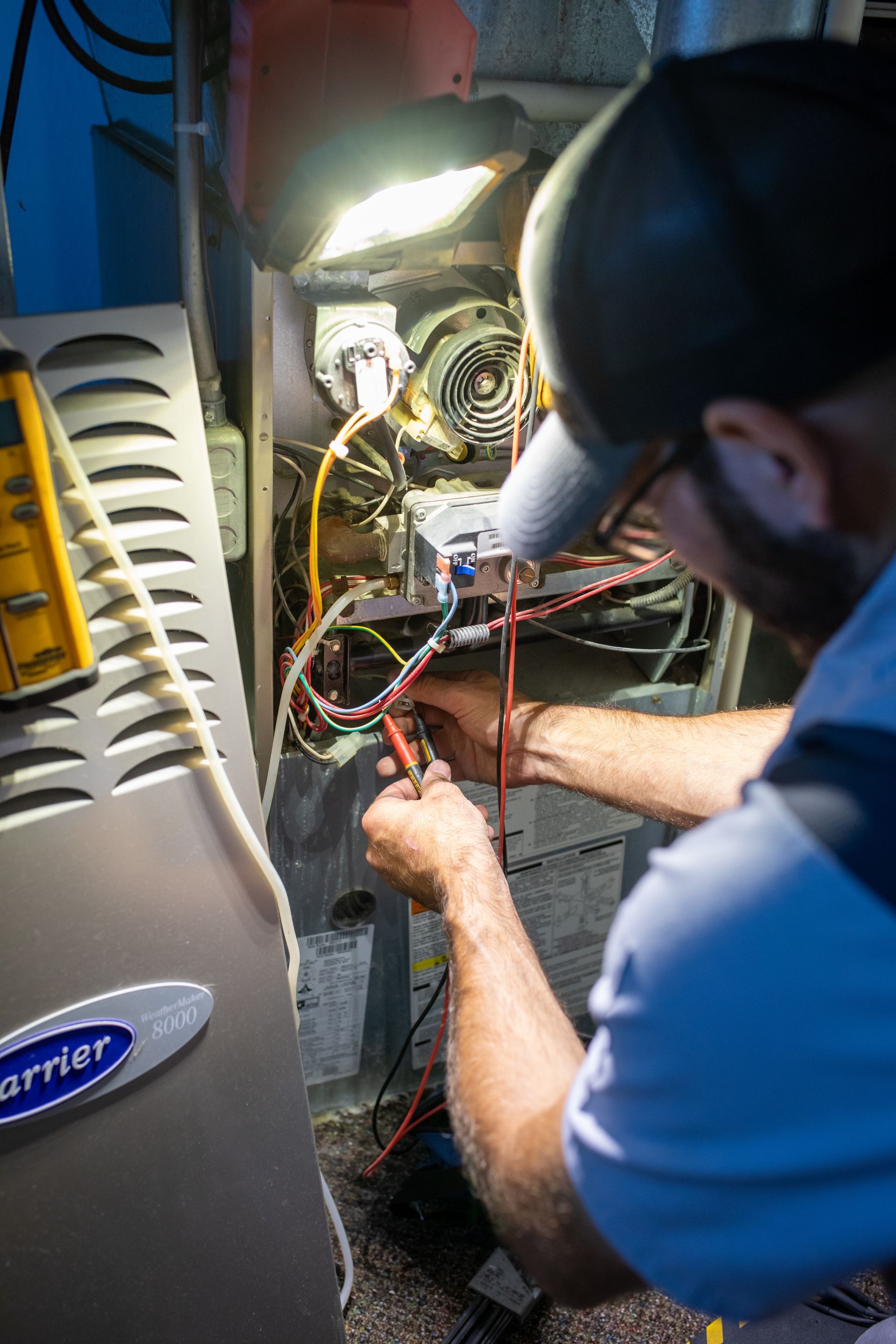 HVAC technician in a cap and glasses repairs a furnace with electrical tools, illuminated by a work light.