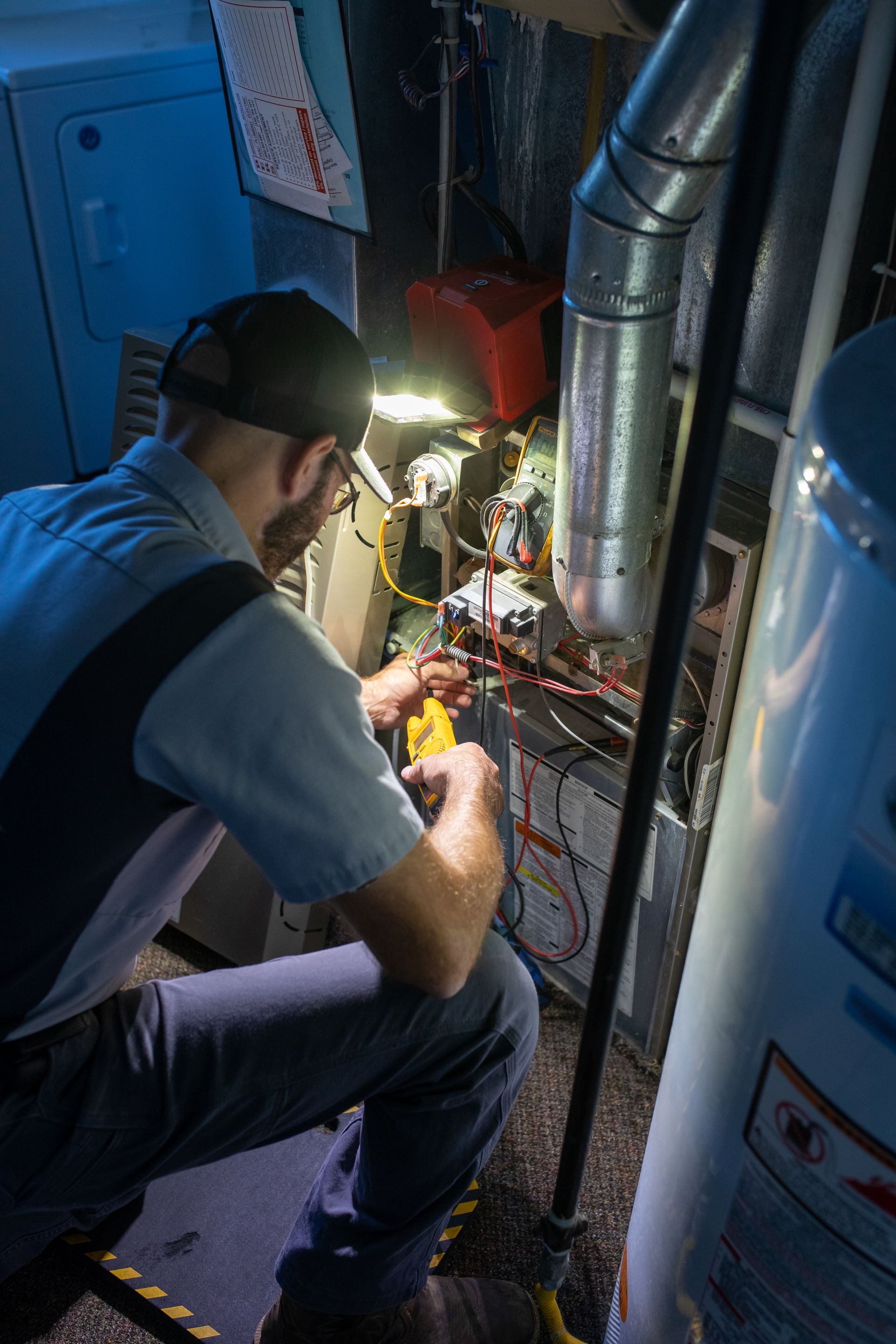 HVAC technician in a utility room, working on a furnace with a tool, lit by a work light.