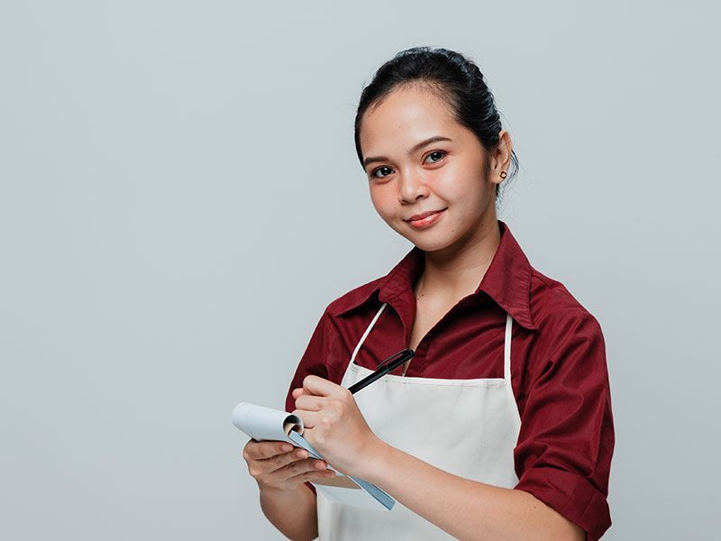 Person in a maroon shirt and apron holding a clipboard and pen against a plain gray background