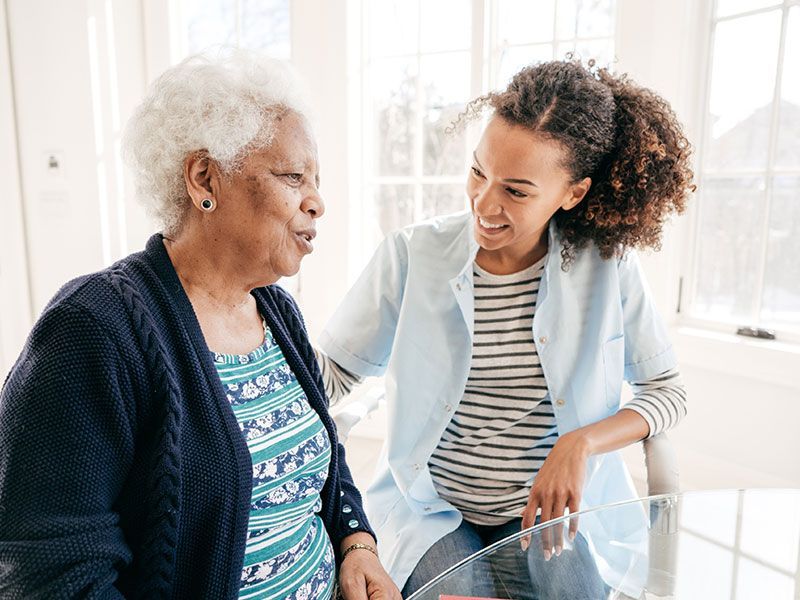 Two women smiling and talking in a bright kitchen, one seated and one leaning beside her.
