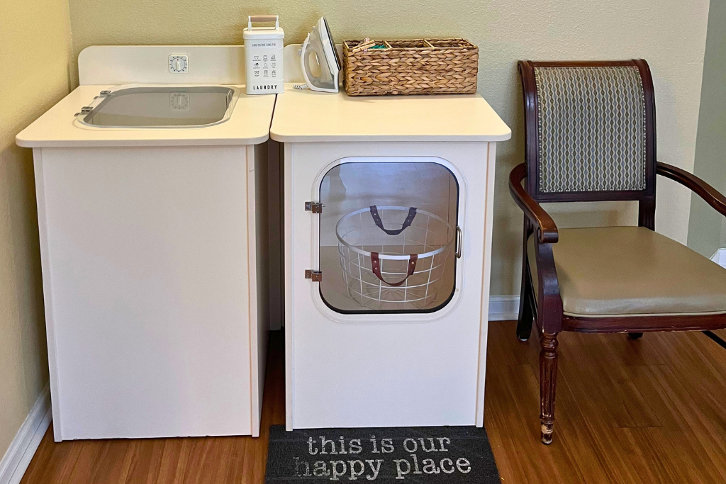 Bathroom vanity with sink, toiletries, pet door, wicker basket, and a chair beside a “this is our happy place” mat