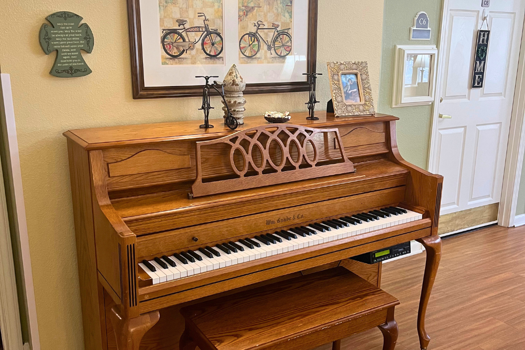 Wooden upright piano in a bright room, with sheet music and framed art on the wall.