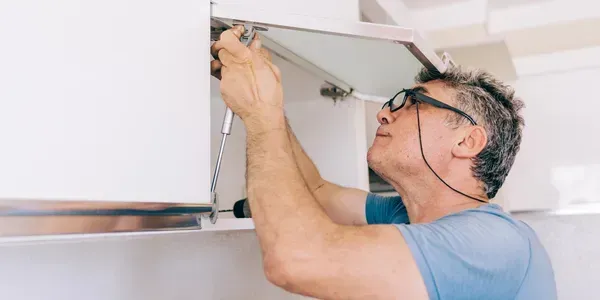 Man in glasses adjusting a white cabinet hinge with a screwdriver in a kitchen