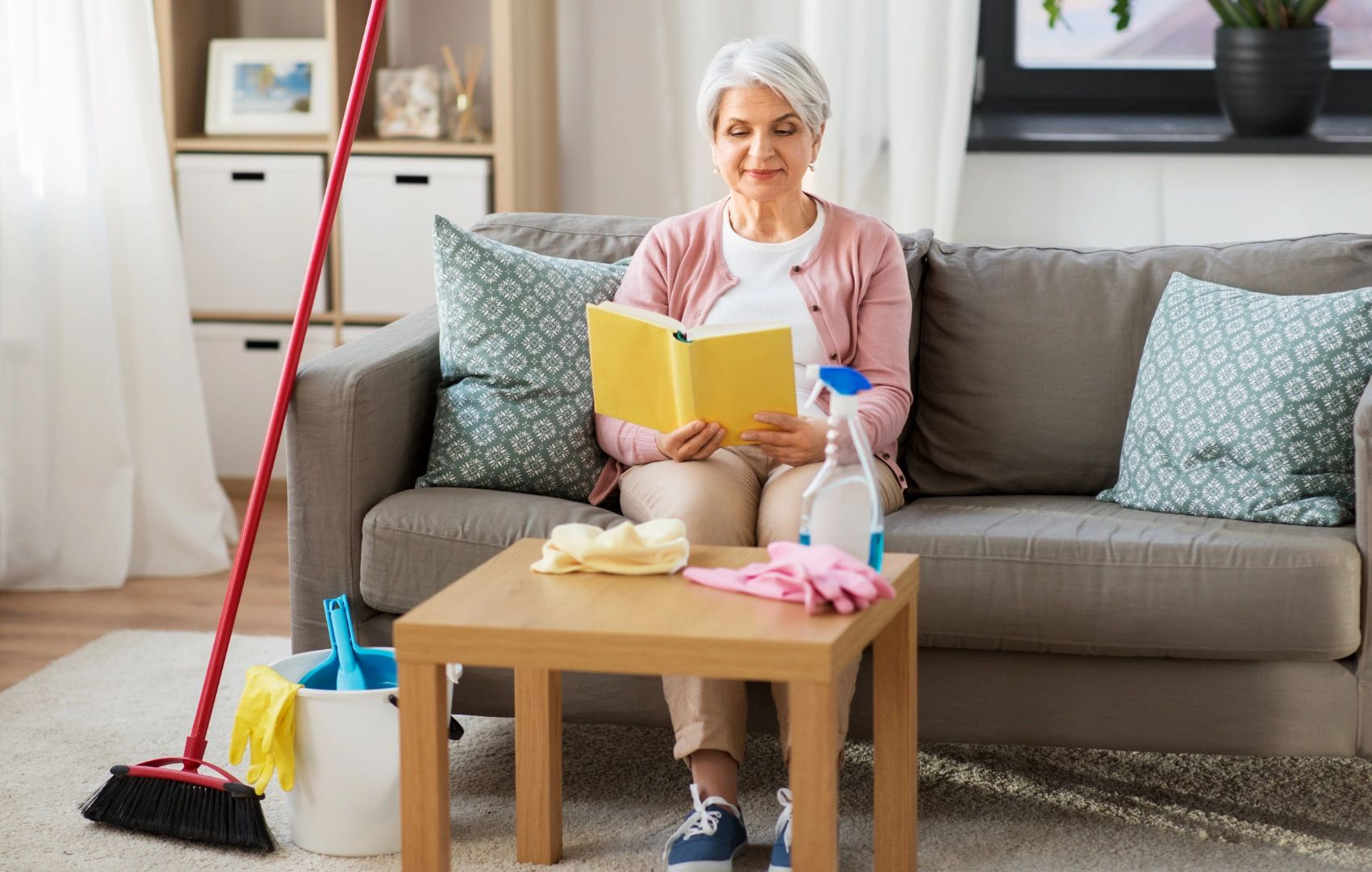 Person sitting on a sofa reading a yellow book in a tidy living room, with a broom nearby.