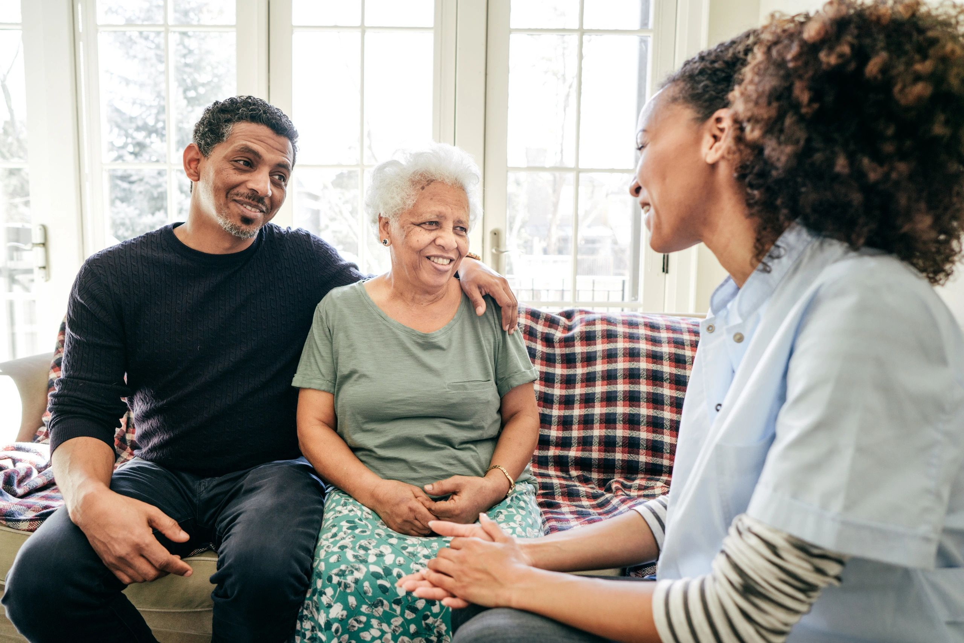 Three people chatting on a couch in a bright living room, smiling and leaning in together.