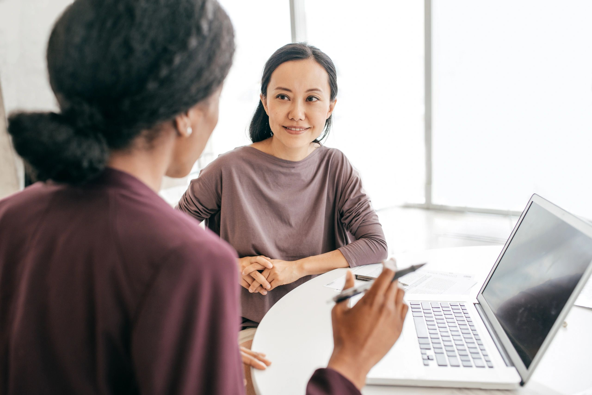 Two people in a bright office meeting across a table with a laptop and notebook