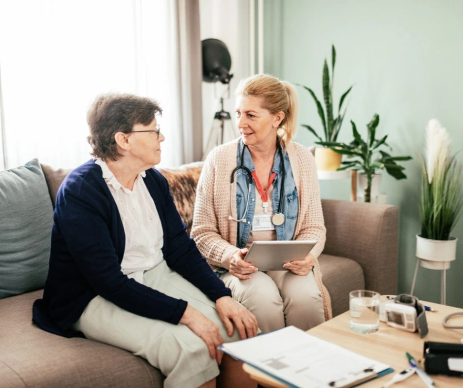 Two people talking on a couch in a bright living room, one holding a tablet.