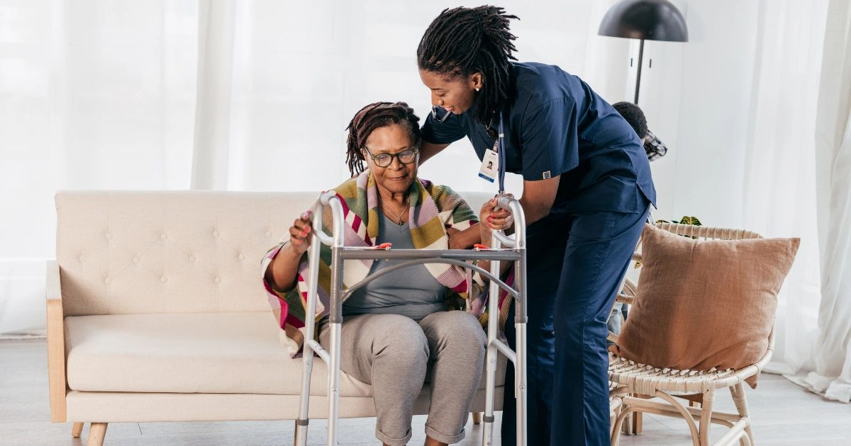 Caregiver assisting a seated woman with a walker in a bright living room