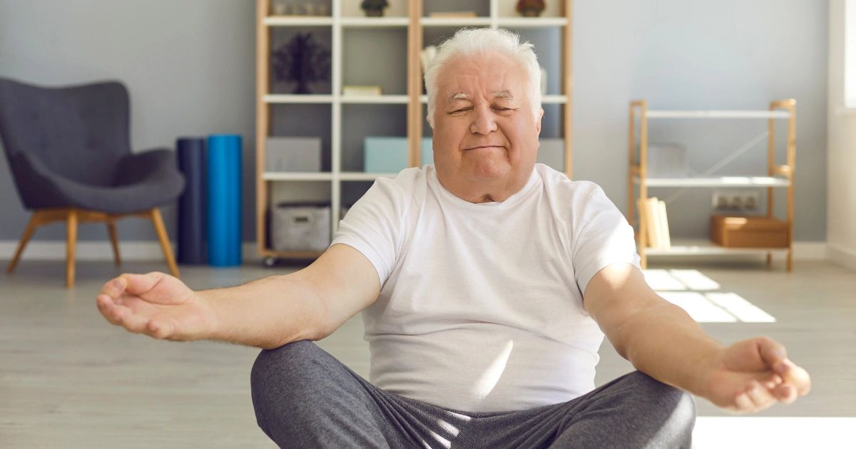 Person sitting cross-legged on a yoga mat, meditating with eyes closed in a bright living room.