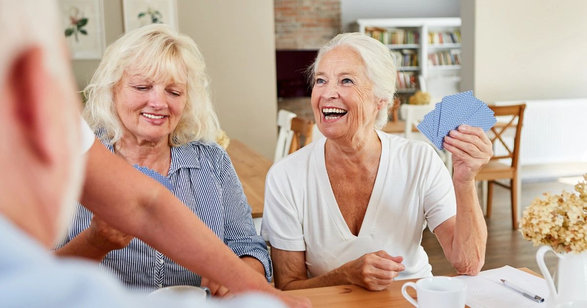 People smiling and chatting at a table, with one holding a blue fan in a bright room