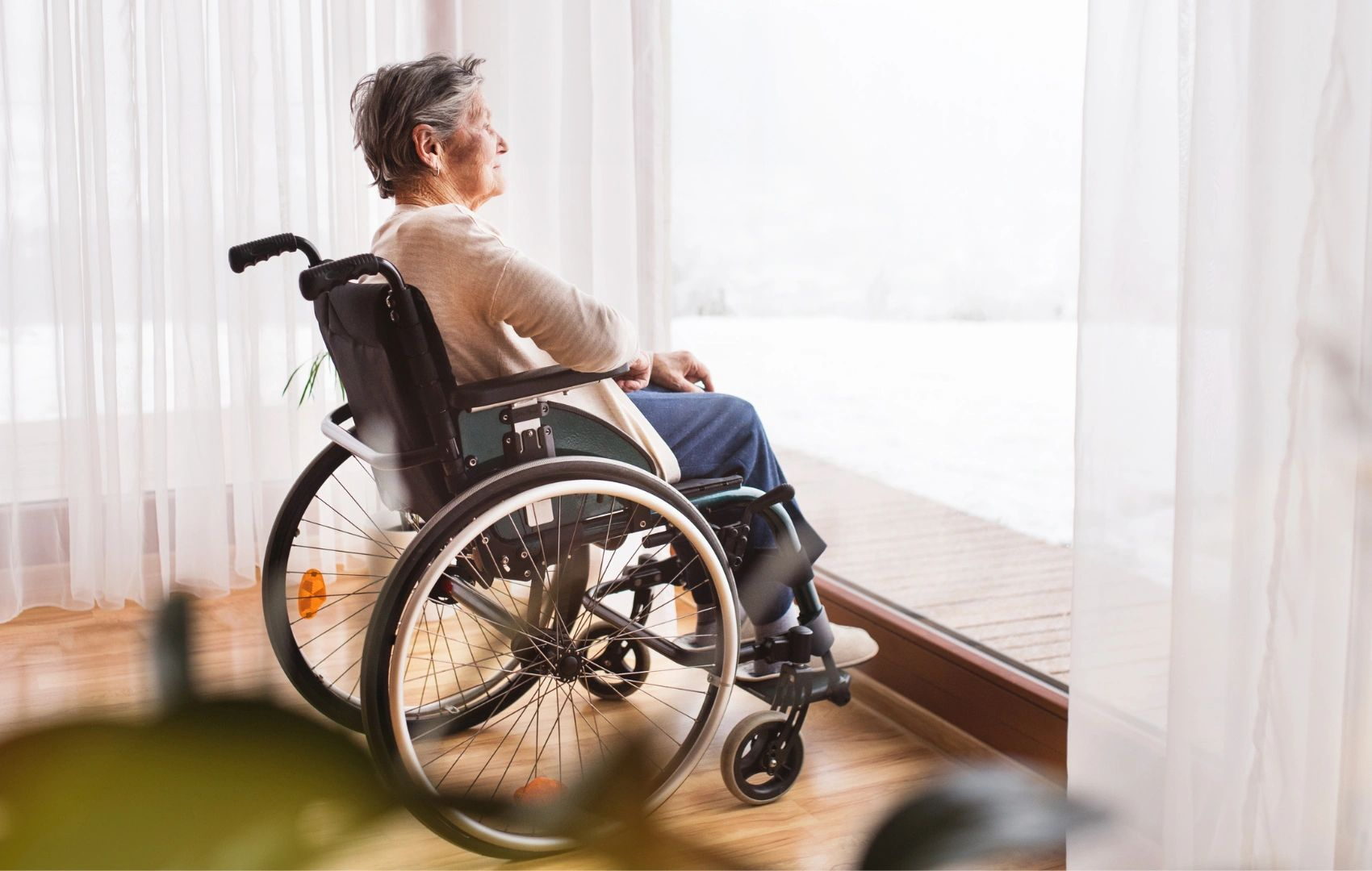 Person in a wheelchair looking out a bright window with sheer curtains