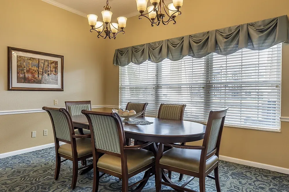 Formal dining room with oval table, six chairs, chandelier, framed art, and large window with blinds and drapes