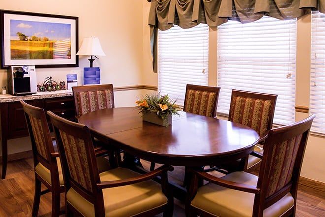 Dining room with oval wooden table, six chairs, and sunlit windows with blinds and drapes.