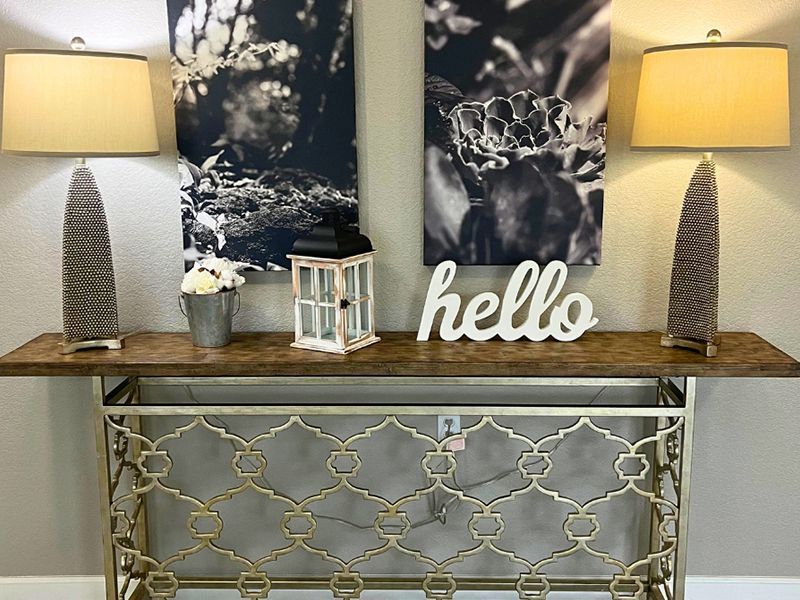 Decorative console table with lamps, floral wall art, a “hello” sign, and gold mirrored cabinet