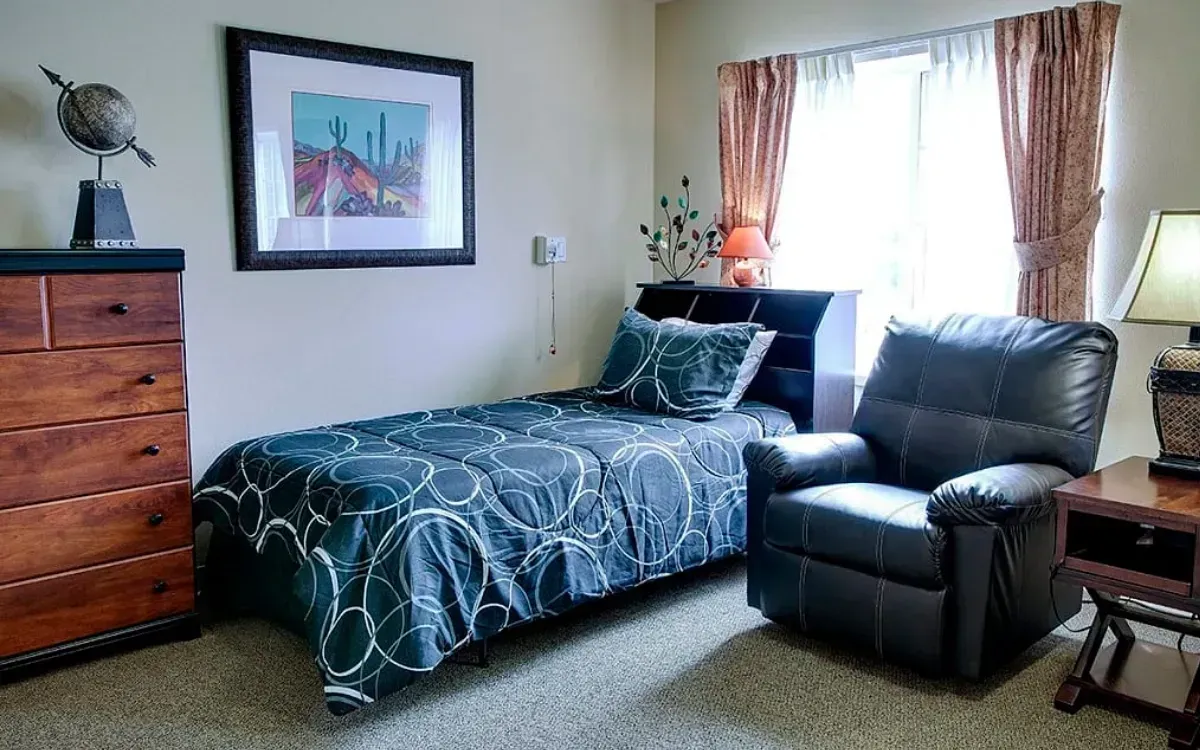 Bedroom with a made bed, gray recliner, wooden dresser, framed art, and window curtains