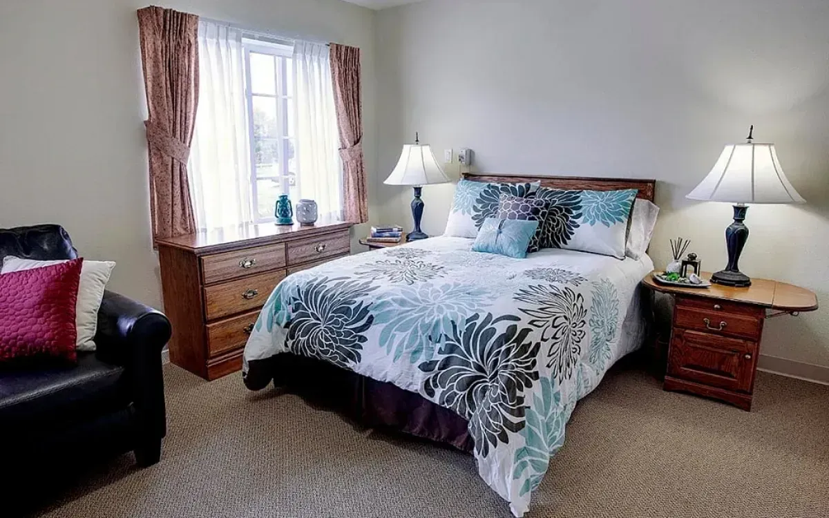 Cozy bedroom with floral bedspread, wooden nightstand lamps, window with curtains, and a black armchair