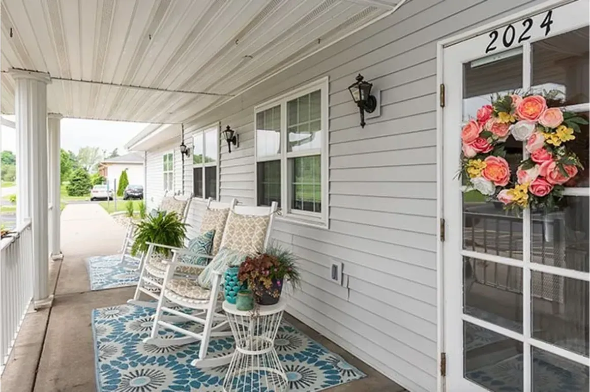 Covered porch with white rocking chairs, blue rug, and a floral wreath on a screened door.