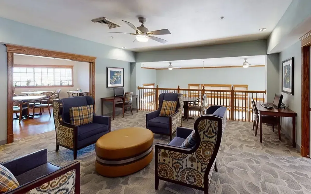 Bright living room with patterned chairs, round ottoman, ceiling fan, and railing overlooking a lower level