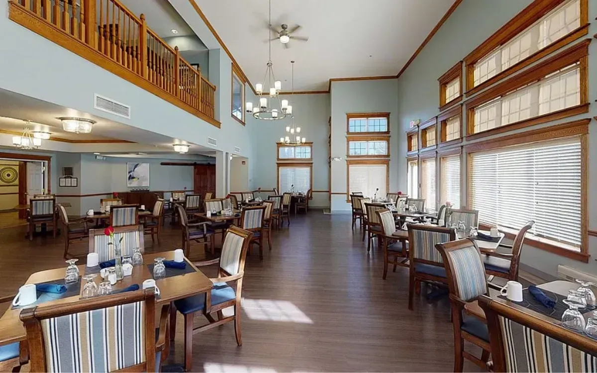 Bright, airy dining hall with wooden tables, chairs, tall windows, and a loft railing above.