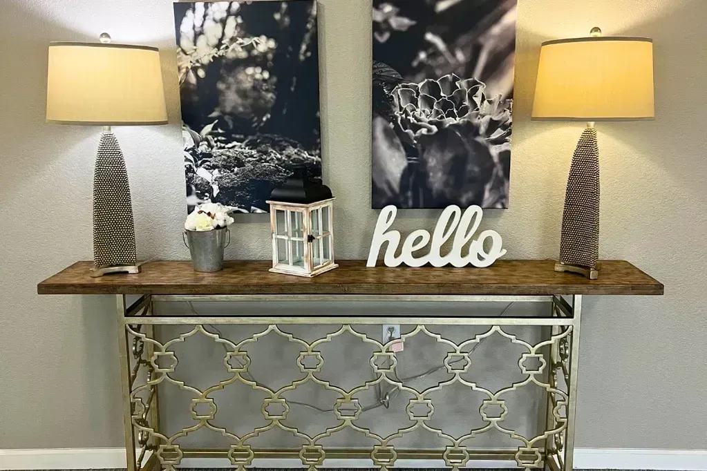 Entryway console table with two lamps, abstract wall art, and a “hello” sign above a gold lattice base