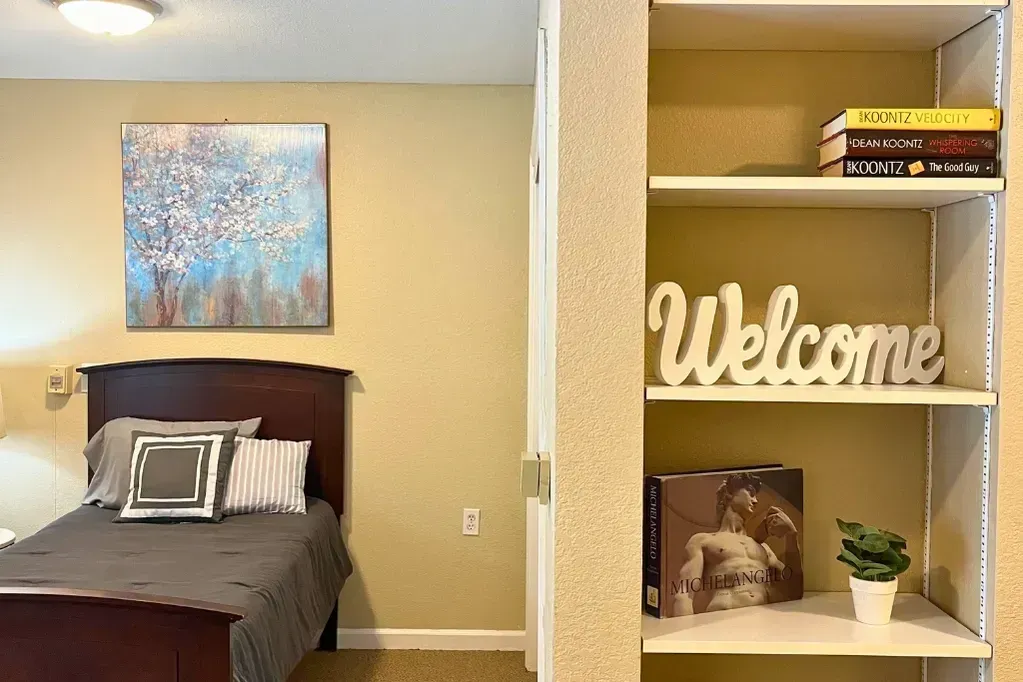 Bedroom with bed, abstract wall art, and bookshelf displaying a “Welcome” sign and books