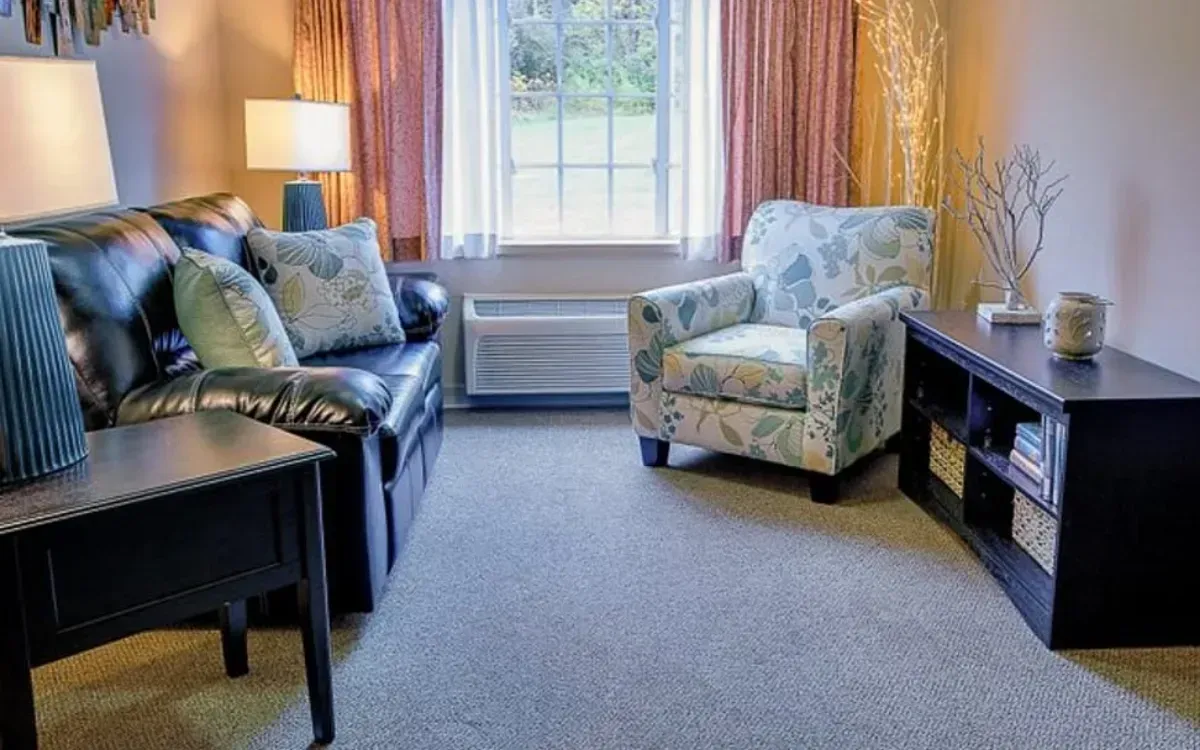 Cozy living room with patterned armchair, dark sofa, side tables, and a window with curtains and lamp.