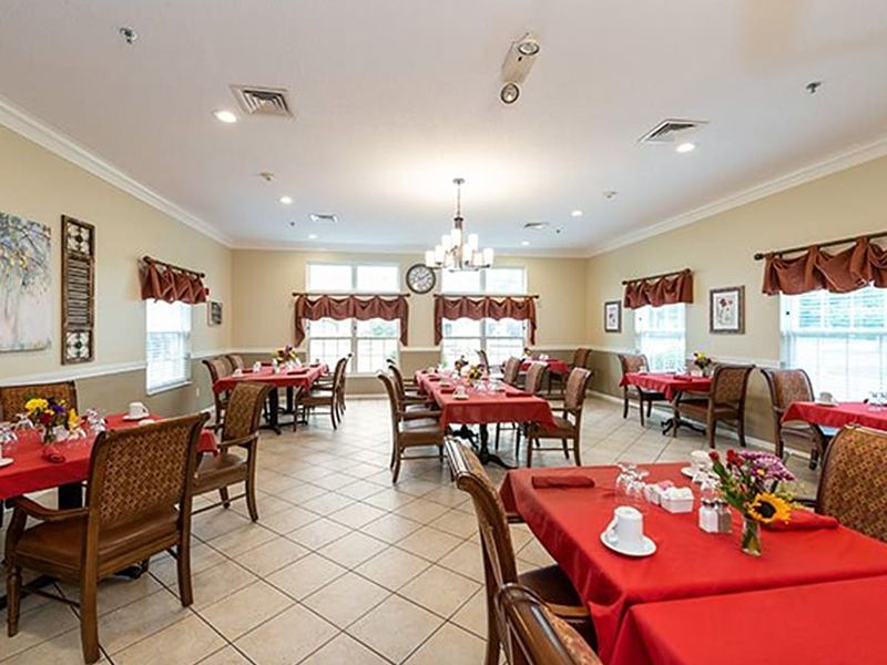 Bright dining room with red tablecloths, wicker chairs, and large windows letting in daylight.
