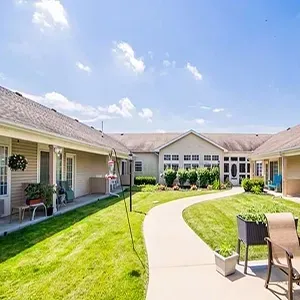 Sunny courtyard with curved walkway, green lawn, and patio chairs between beige apartment buildings