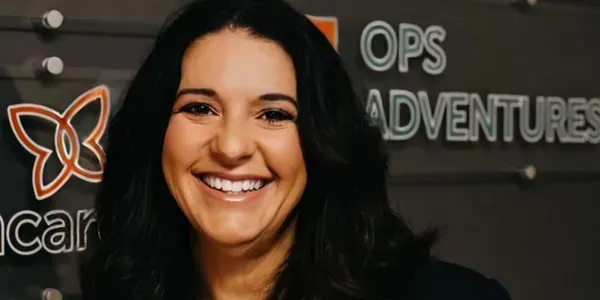 Smiling woman in black against a dark sign reading “Ops Adventures”