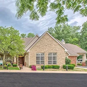 Single-story tan brick house with gabled roof, front porch, and green trees lining the street