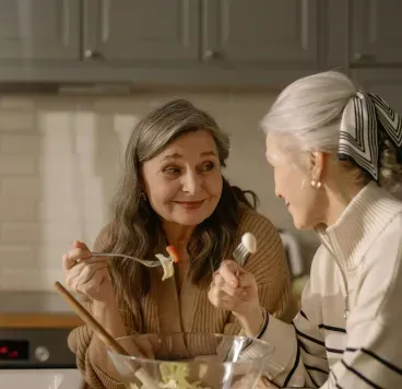 Two people talking over salad in a kitchen, with one holding a fork and a bowl on the counter.