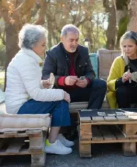Three people sitting on pallet benches outdoors, chatting with cups in a sunny park.
