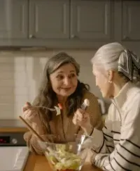 Two people in a kitchen talking while preparing a salad at the counter.