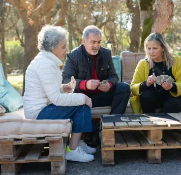 Three people sit on pallet benches outdoors, chatting around a small table in a wooded park.